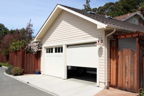Garage résidentiel avec porte sectionnelle blanche partiellement ouverte, façade en bardage bois peint beige, toiture à pignon avec bardeaux d'asphalte